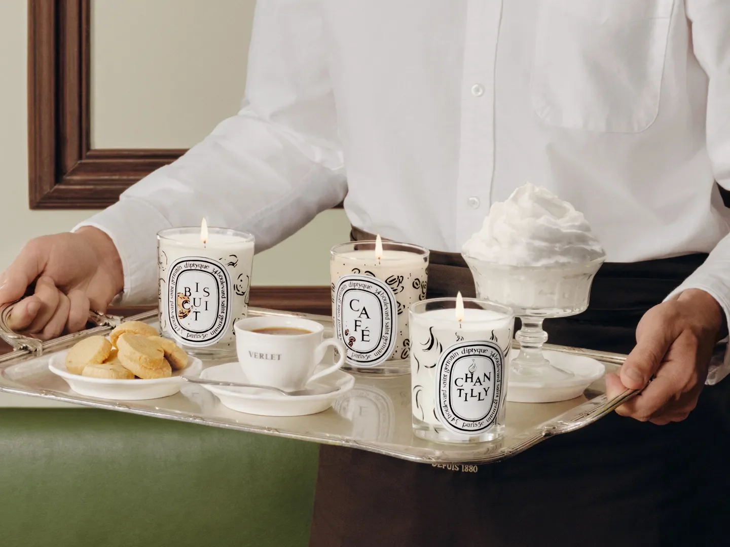 A man carrying a tray with coffee, cream, cookies, and three Diptyque candles, setting the perfect atmosphere